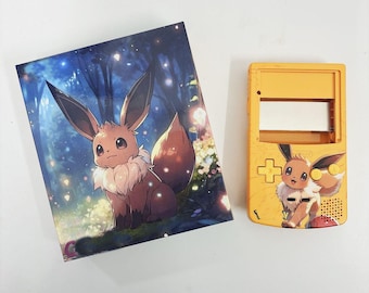 Puede incluir: Una consola de videojuegos portátil amarilla y marrón con un personaje Eevee marrón en la pantalla. La consola tiene un botón rojo y un botón amarillo. La consola está sobre una superficie blanca.