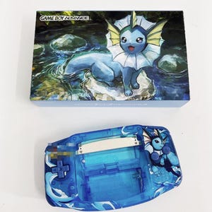 May include: A blue Game Boy Advance handheld gaming console with a custom design featuring a blue water-type Pokemon character,  "GAME BOY ADVANCE" is printed on the box.