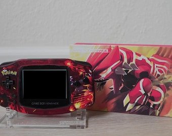 May include: A red and black Game Boy Advance with a Pokemon design. The console has a black screen and the words "Game Boy Advance" are printed on the bottom. The console is sitting on a clear stand. The box art features a red and yellow Pokemon character.