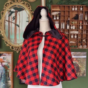 May include: A red and black plaid hooded cape displayed on a mannequin. The cape has a black satin lining and a decorative clasp closure. The cape is a classic design, perfect for cosplay or costume events.