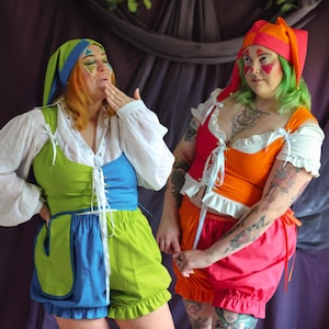 May include: Two individuals wearing colorful jester-inspired outfits. One wears a green and blue bodice with matching shorts and a green cap. The other wears a pink and orange bodice with matching shorts and a pink and orange jester hat.
