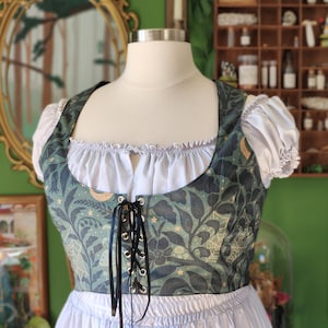 May include: A green and blue patterned bodice with black lacing, worn with a white blouse and skirt. The bodice has a botanical design with moons and stars. The outfit is displayed on a mannequin.