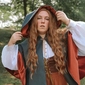 May include: A woman wearing a hooded cloak with a dark green outer layer and a burgundy lining. The cloak has a long, flowing design and is worn over a white blouse and a brown skirt. The woman has long, curly red hair and is looking at the camera.