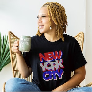 May include: A person wearing a black t-shirt with the words "NEW YORK CITY" in red, white, and blue. The person is holding a green mug with a dragonfly design.