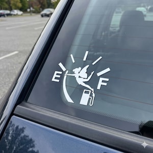 May include: A white vinyl decal applied to a car window featuring a fuel gauge design. The graphic shows a silhouette of a fairy figure positioned between the letters E and F, with a fuel pump icon at the base of the gauge needle indicator.