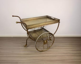 May include: A gold-toned metal serving cart featuring two rectangular tiers with mesh surfaces. The cart has a curved handle, two large decorative spoked wheels, and two smaller swivel casters at the base for mobility. The frame is constructed from thin metal tubing.