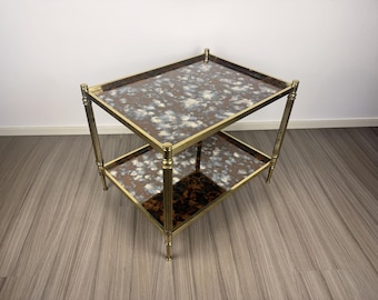 May include: A two-tiered side table with a gold-colored metal frame. The table has rectangular shelves with a tortoiseshell pattern. The top shelf has a speckled design in brown, white, and blue. The table is approximately 24 inches long and 16 inches wide.