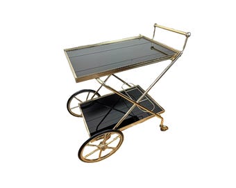 Op de afbeelding: Een gouden metalen serveerwagen met twee zwarte glazen planken en wielen. De wagen heeft een handvat aan de bovenkant.