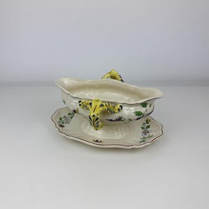 May include: A cream-coloured ceramic gravy boat with an attached underplate, featuring yellow handles with blue accents and a hand-painted floral pattern in green, blue, and brown. The piece has a scalloped rim and a moulded, ribbed texture.