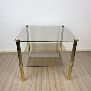 Puede incluir: Una mesa auxiliar cuadrada de cristal con dos estantes de cristal. La mesa tiene cuatro patas doradas, estriadas, con detalles dorados en la parte superior e inferior. La mesa mide aproximadamente 61 cm de ancho y 61 cm de profundidad.