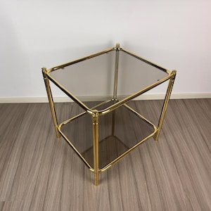 Puede incluir: Una mesa auxiliar cuadrada de dos niveles con estructura de metal dorado y dos superficies de vidrio ahumado. La mesa tiene un diseño clásico con forma geométrica, ideal para salón o dormitorio. La mesa mide aproximadamente 51 cm de largo, ancho y alto.