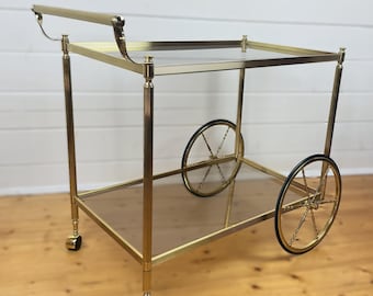 Op de afbeelding: Een goudkleurige bartrolley met twee heldere glazen planken en een handvat. De kar heeft vier kleine wielen en twee grotere spaakwielen met zwarte rubberen banden. De kar staat op een houten vloer.
