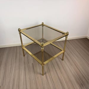 May include: A square, two-tiered side table with a brass-colored metal frame and glass shelves. The table has a classic design with rounded finials at the top of each leg. The table is approximately 20 inches wide and 20 inches long.