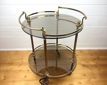 May include: A two-tiered brass serving cart with smoked glass shelves and a brass handrail. The cart has four wheels and is on a wooden floor.