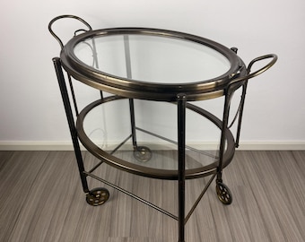 May include: An antique-style serving cart with two oval glass shelves and a bronze-colored metal frame. The cart has small wheels for easy movement and curved handles on each side. A stylish piece for entertaining.