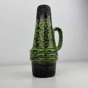 May include: A green and black ceramic vase with a handle. The vase has a textured, speckled pattern in shades of green and black. The base and top rim are a rough, dark black texture. The vase is approximately 25 cm tall.