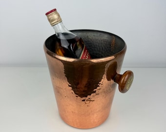 May include: A copper ice bucket with a hammered finish and a wooden handle. A small bottle of liquor is inside the bucket. The bucket has a wide opening and a tapered shape.