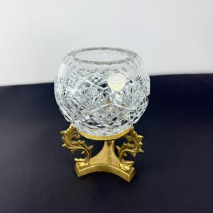 Pu&ograve; includere: Una ciotola di cristallo trasparente tagliato con forma rotonda, appoggiata su un supporto decorativo in metallo color oro. La ciotola presenta intricati motivi geometrici e un piccolo adesivo rotondo. Il supporto ha una base curva e motivi a foglie stilizzate.