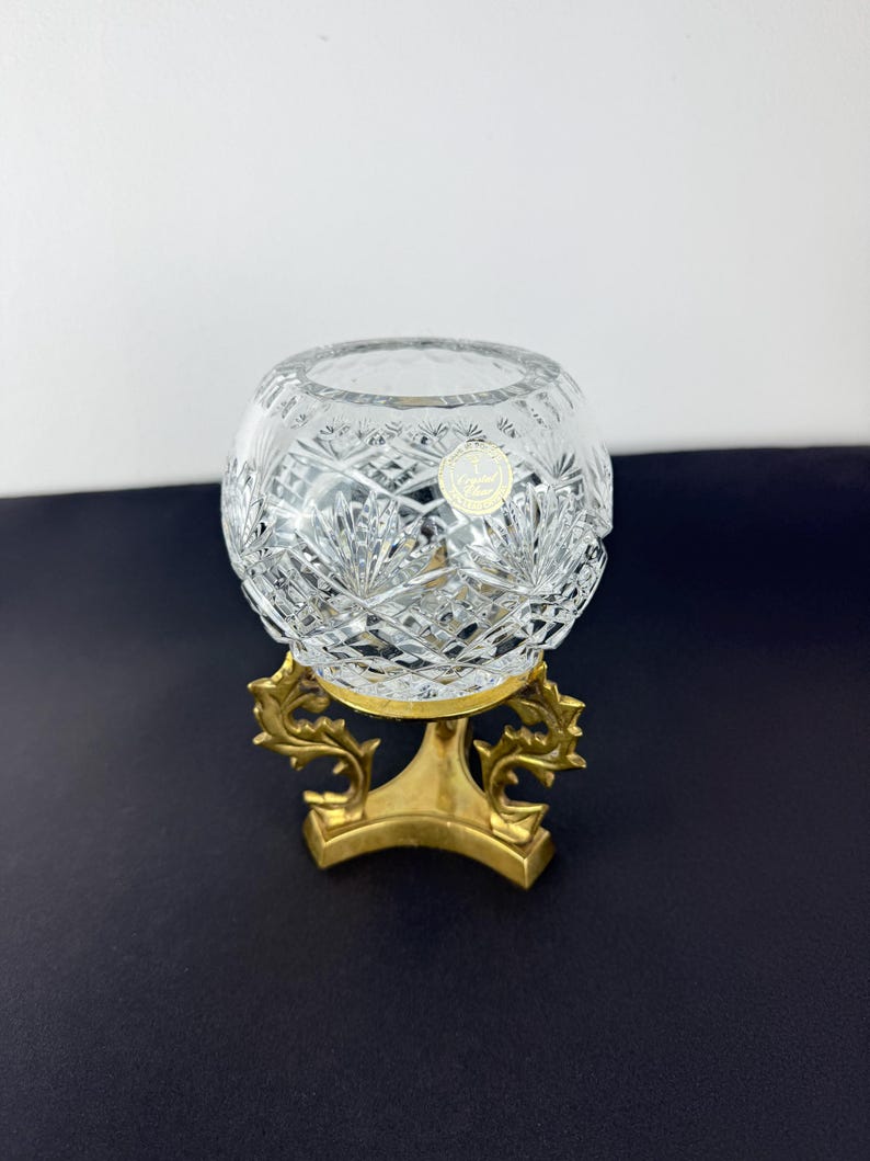 Pu&ograve; includere: Una ciotola di cristallo trasparente tagliato con forma rotonda, appoggiata su un supporto decorativo in metallo color oro. La ciotola presenta intricati motivi geometrici e un piccolo adesivo rotondo. Il supporto ha una base curva e motivi a foglie stilizzate.