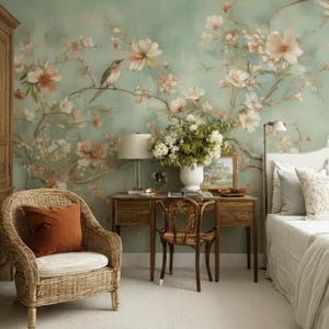 Puede incluir: Un dormitorio con un mural de pared verde salvia con ramas florales pintadas en blanco y rosa y un pequeño pájaro. La habitación incluye un sillón de mimbre con un cojín color óxido, un escritorio de madera con lámpara, un florero y una cama.