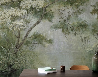 Puede incluir: Un mural de pared con una escena de bosque pintada con árboles de flores blancas, follaje verde y un cuerpo de agua tranquilo. En primer plano, una mesa de madera oscura sostiene una pila de libros blancos, una taza marrón y un jarrón de vidrio verde.