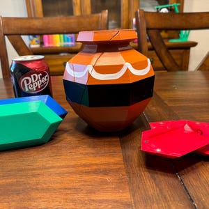 May include: A brown and black ceramic pot with white accents, a green and blue geometric shape, and a red geometric shape. The pot is a puzzle and has a design that resembles a traditional pot. The objects are on a wooden table.