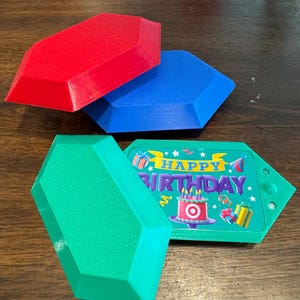Puede incluir: Tres formas geométricas impresas en 3D en rojo, azul y verde. La forma verde tiene una tarjeta de cumpleaños adjunta con el texto "Feliz cumpleaños" y un logotipo de Target.