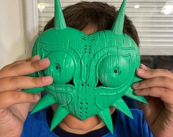 Puede incluir: Una máscara de plástico verde en forma de corazón con puntas afiladas. La máscara tiene dos grandes agujeros para los ojos y un agujero más pequeño en el centro.