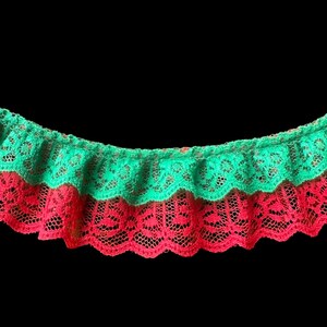 May include: Close-up of two layers of decorative lace trim. The top layer is a vibrant green, and the bottom layer is a bright red. Both layers have a scalloped edge and a delicate, openwork pattern, set against a black background.
