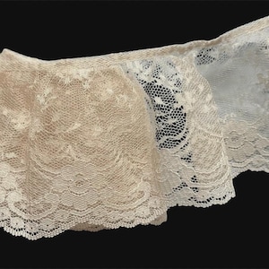 May include: A length of ruffled lace trim featuring a delicate floral pattern. The fabric displays a gradient of color, transitioning from a warm beige or tan tone on the left side to a crisp, bright white on the right side, with scalloped edges.