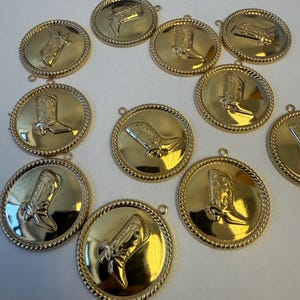May include: Gold-colored round pendants with a raised image of a cowboy boot. Each pendant has a rope-like border and a small loop at the top for attaching to a chain or other jewelry. The pendants are likely for jewelry making.