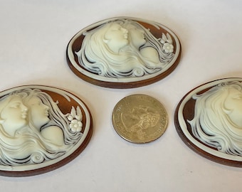 May include: Three oval-shaped cameo pendants with a brown background and white raised design of two women's faces with flowing hair. The design is reminiscent of Art Nouveau style.