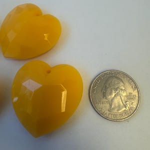 May include: Two faceted, yellow heart-shaped beads with a hole at the top. A U.S. quarter coin is next to the beads for size comparison. The beads have a glossy finish and are likely made of plastic or resin.