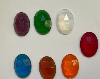 Könnte beinhalten: Sortiment aus sieben ovalen, facettierten Glasperlen in verschiedenen Farben, darunter Lila, Grün, Weiß, Rot, Blau und Orange. Die Perlen sind in einer Reihe auf einer weißen Oberfläche angeordnet.