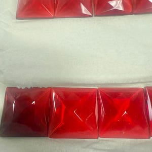 May include: Rows of square, faceted, red glass beads. The beads are arranged in a line, with a slight variation in color and clarity. The beads are likely for jewelry making or other craft projects.