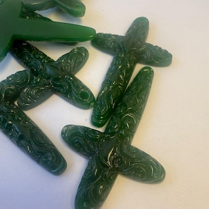 May include: A collection of dark green, cross-shaped beads with a glossy finish. Each bead features an intricate, swirling pattern carved into the surface. The beads are scattered across a white surface, showcasing their detailed design.