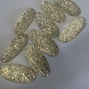 May include: A collection of nine oval-shaped cabochons featuring a clear, textured surface with a multifaceted, crushed-glass appearance. Each piece has a smooth, rounded edge and a reflective, shimmering quality that catches light from multiple angles.