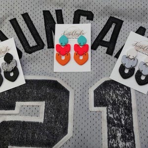 May include: Three pairs of handmade polymer clay earrings in various colors. One pair is black and silver glitter, another is orange, red, and teal, and the third is black, gray, and white. Each pair is displayed on a white card with the text "Frestillagla".