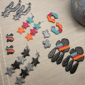 May include: A collection of handmade earrings in various designs. Styles include silver glitter cactus shapes, star clusters in orange, pink, and turquoise, and abstract shapes with color blocks. Other earrings are black with glitter.
