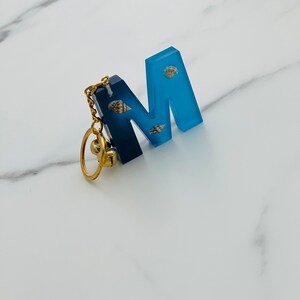 Può includere: Un portachiavi in resina blu a forma di lettera M con una catena dorata e un piccolo ciondolo dorato. La lettera M ha piccole conchiglie incastonate nella resina.