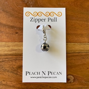 May include: A soccer ball zipper pull with a silver-tone metal chain and clasp. The soccer ball charm is black and silver. The product is displayed on a white card with the text "Zipper Pull" and "PEACH N PECAN".