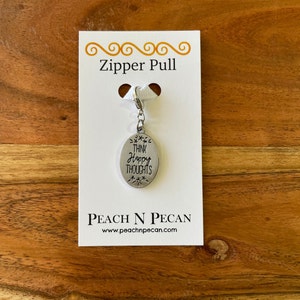 Pode incluir: Um puxador de zíper oval prateado com as palavras "THINK HAPPY THOUGHTS" gravadas. O puxador está preso a um cartão branco com as palavras "Zipper Pull" e o nome da marca "PEACH N PECAN". O fundo é uma superfície de madeira.