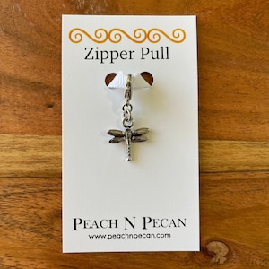 May include: Silver dragonfly zipper pull charm with a white ribbon and the text "Zipper Pull" and "Peach N Pecan" with the website address "www.peachnpecan.com".