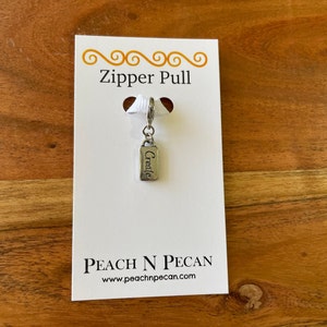以下が含まれることがあります： 「Create」と刻まれた銀製のジッパーの引き手。ジッパーの引き手は、「Zipper Pull」と「Peach N Pecan」の文字とウェブサイトのアドレス「www.peachnpecan.com」が書かれた白いカードの上に置かれています。