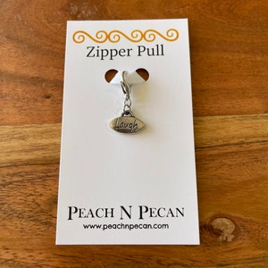 Pode incluir: Puxador de zíper de prata com a palavra "Laugh" em um charme oval. O puxador está em um cartão branco com o texto "Zipper Pull" e "Peach N Pecan" com o endereço do site.