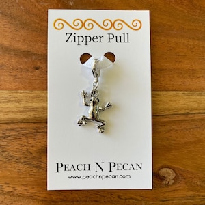 Könnte beinhalten: Silberner Frosch-Reißverschlussanhänger mit einem weißen Band. Der Anhänger befindet sich auf einer weißen Karte mit dem Text "Zipper Pull" und "PEACH N PECAN" sowie der Website-Adresse "www.peachnpecan.com".