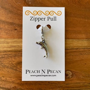 May include: A silver dolphin zipper pull with a silver clasp. The zipper pull is on a white card with the text "Zipper Pull" and "Peach N Pecan" with the website address "www.peachnpecan.com".