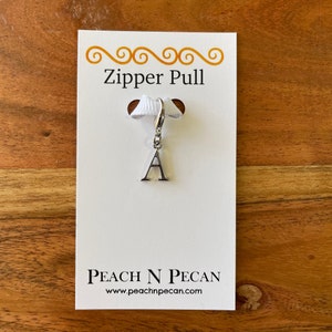 Puede incluir: Tirador de cremallera de plata con la letra A en una tarjeta blanca con el texto "Zipper Pull" y "Peach N Pecan" con la dirección del sitio web "www.peachnpecan.com".