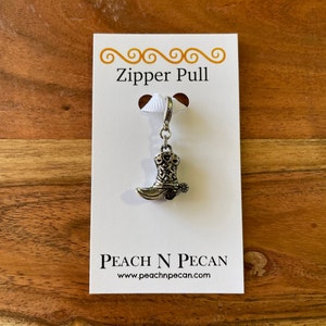 May include: A silver zipper pull with a boot charm. The boot is detailed with a western design and has a spur. The card reads "Zipper Pull" and "Peach N Pecan" with the website address.