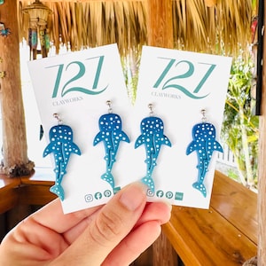 May include: Two pairs of blue whale shark earrings displayed on white 727 Clayworks branded cards. Each earring features a blue shark shape with white polka dot patterns and a silver-toned metal hook. The cards include social media icons.